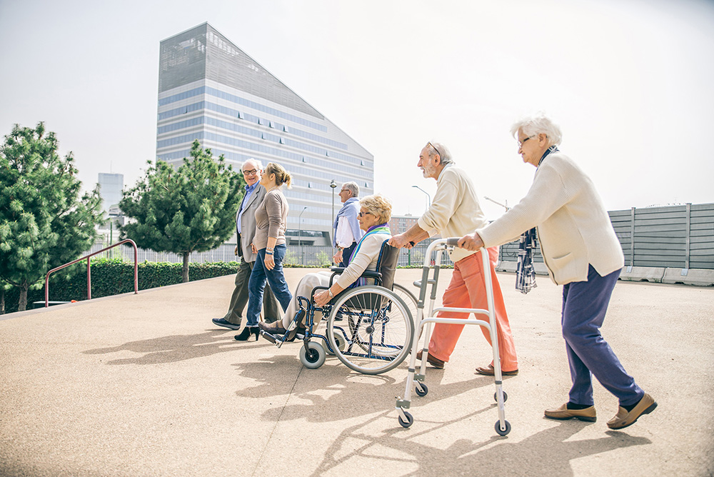 Eine Gruppe von alten Menschen gehen mit Hilfe von Rollstuhl und Rollator auf Spaziergang